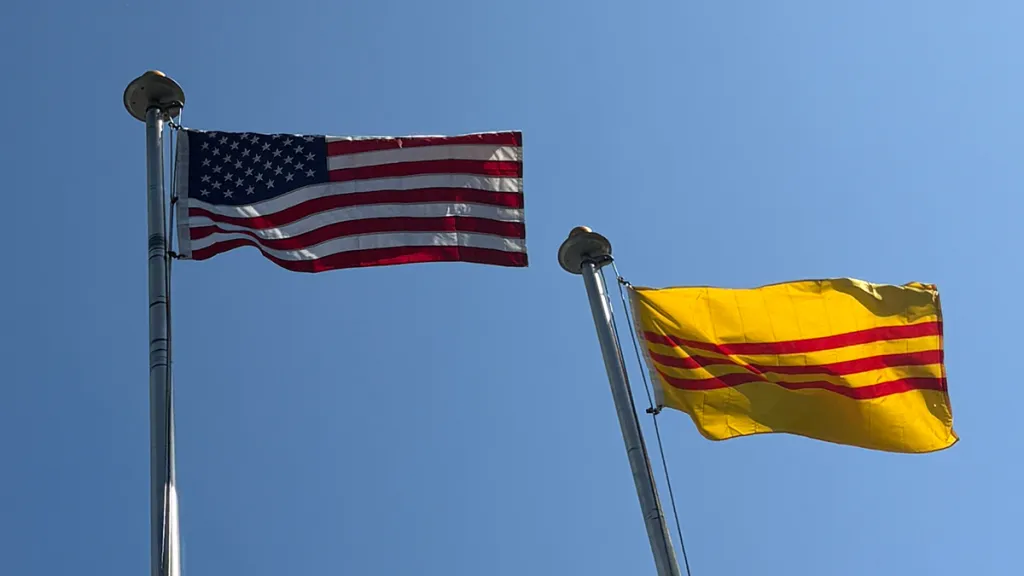 Here’s a concise alt-text suggestion under 100 characters: **"U.S. flag and yellow flag with red stripes flying on flagpoles against blue sky."** Would you like me to also create versions that name the second flag explicitly?