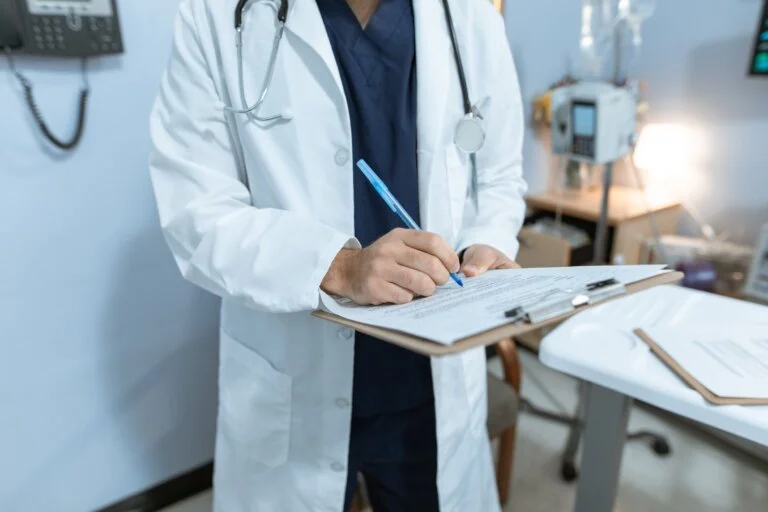 A doctor in a white coat with a stethoscope writes on a patient's chart on a clipboard.