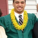 A smiling young man celebrates his graduation wearing a green gown and a yellow lei.