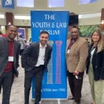 A group of four attendees poses next to "The Youth & Law Forum" event banner.