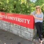 A young woman with blonde hair and sunglasses poses next to the Seattle University sign.