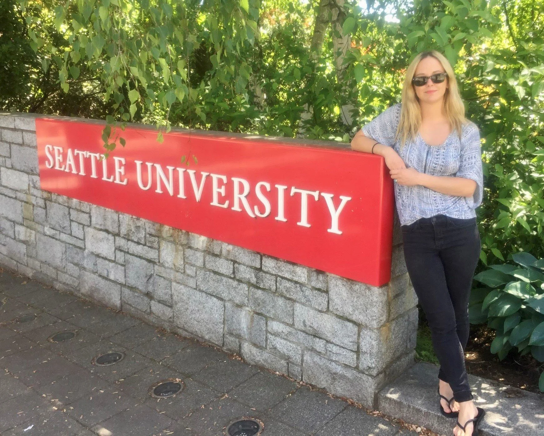 A young woman with blonde hair and sunglasses poses next to the Seattle University sign.