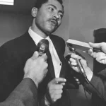 Black-and-white photo of a man in a suit speaking to several reporters holding microphones.