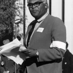 Man in suit speaking at microphone while holding papers outdoors.