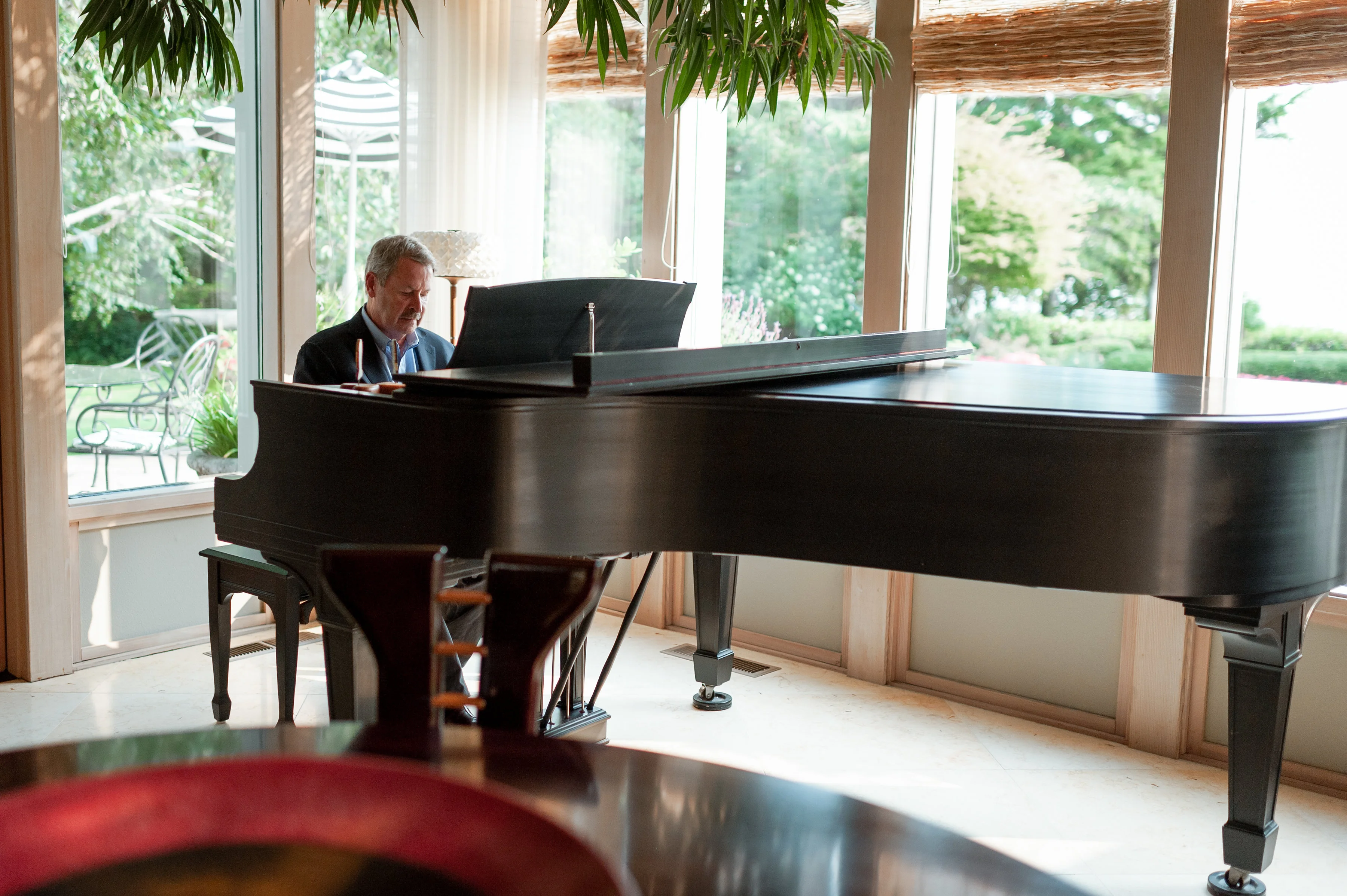 A man in a suit plays a black grand piano in a sunlit room with large windows.