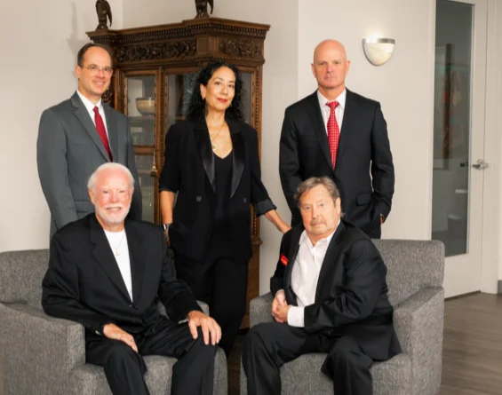 Group portrait of five professionals in business attire, with two seated on chairs and three standing behind them in an office setting. attorneys