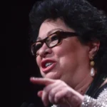 "Woman in glasses speaking on stage, wearing pearl earrings and sparkling attire"