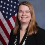 "Smiling woman in professional attire standing in front of an American flag backdrop"