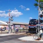 "Amtrak train passing a railroad crossing with cyclists and pedestrians waiting nearby"