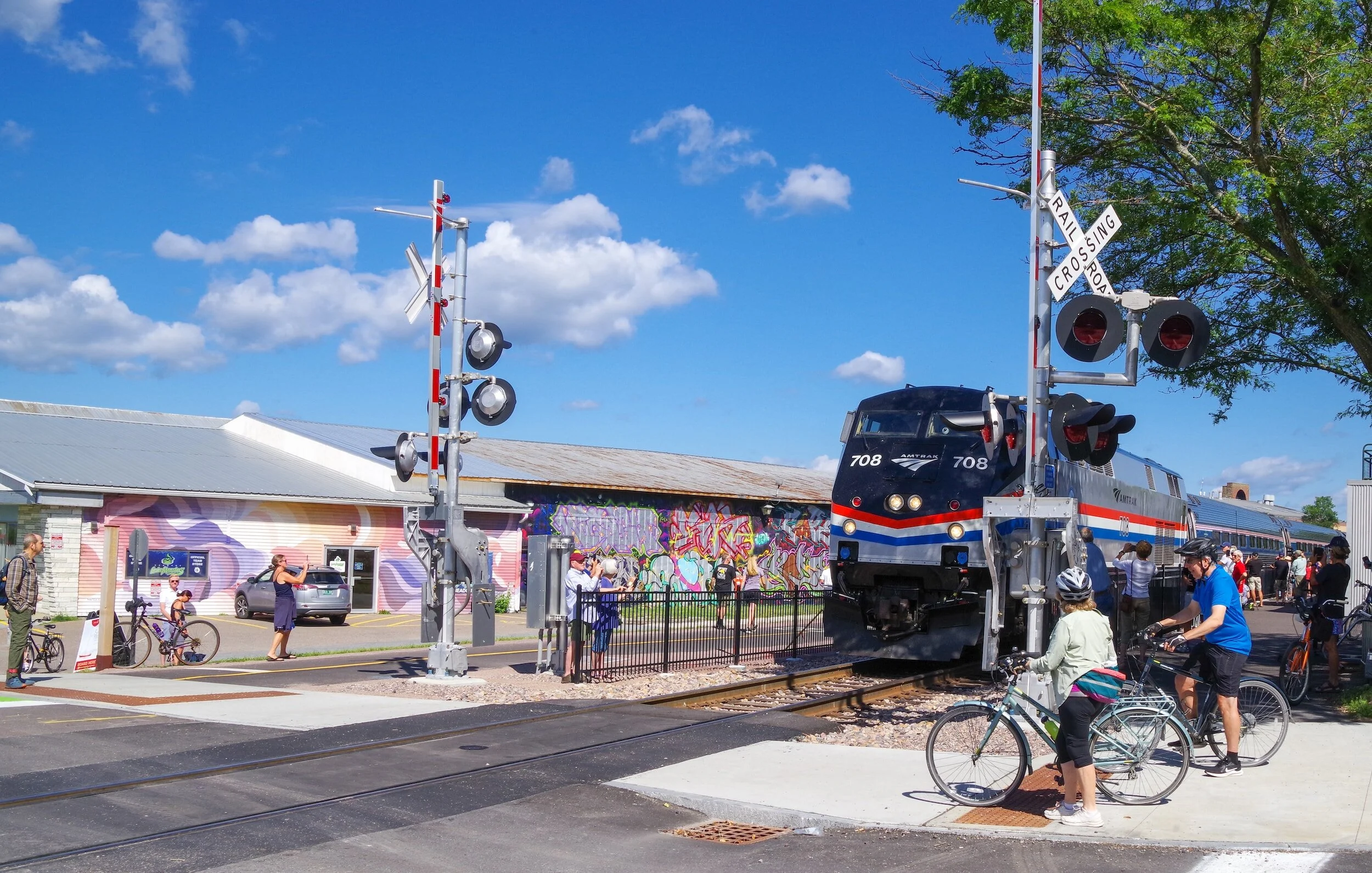 "Amtrak train passing a railroad crossing with cyclists and pedestrians waiting nearby"