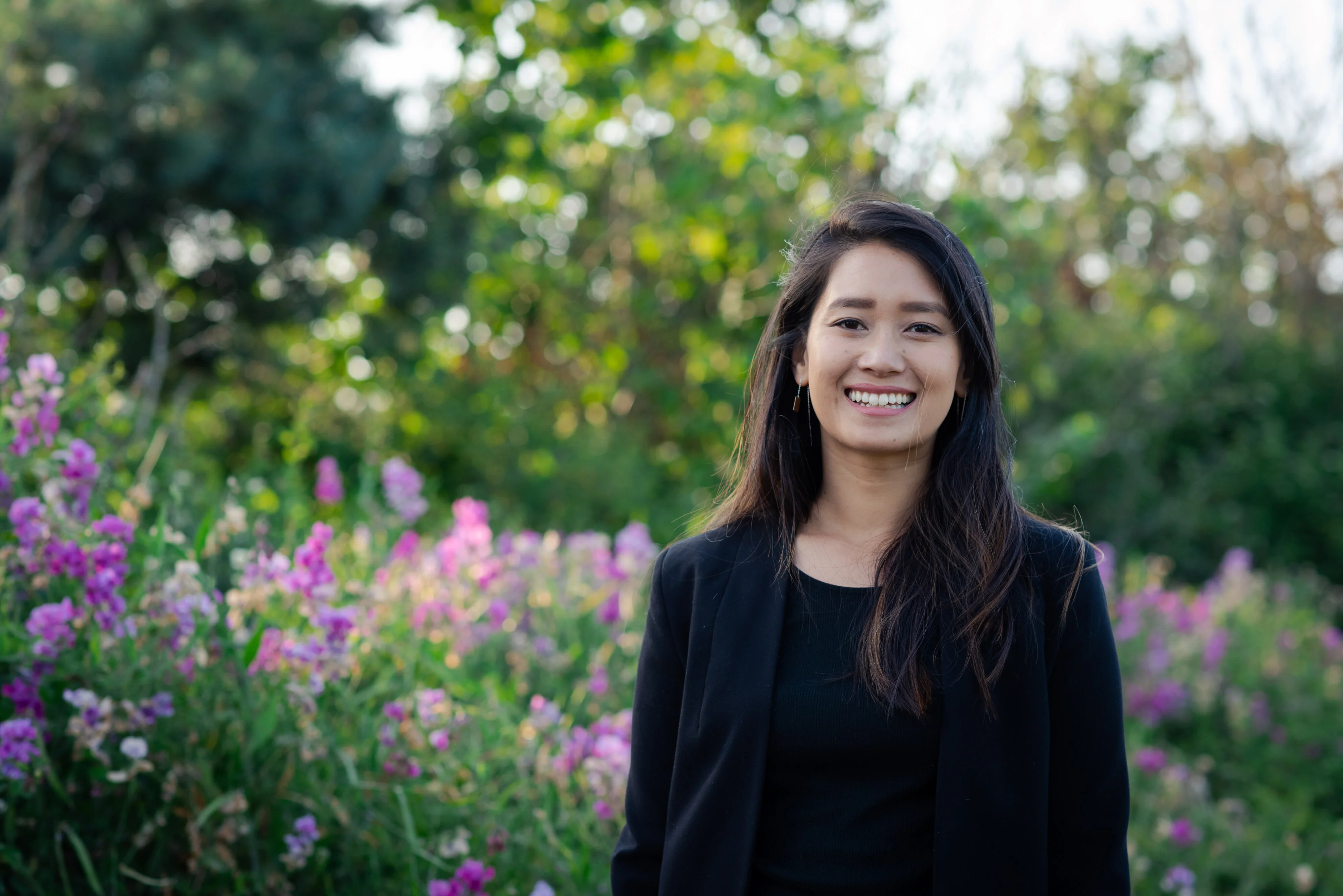 An outdoor portrait of a smiling woman with long dark hair, in front of a field of flowers.