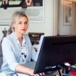 A blonde woman in a light blue blazer sits at her office desk and looks at the camera.