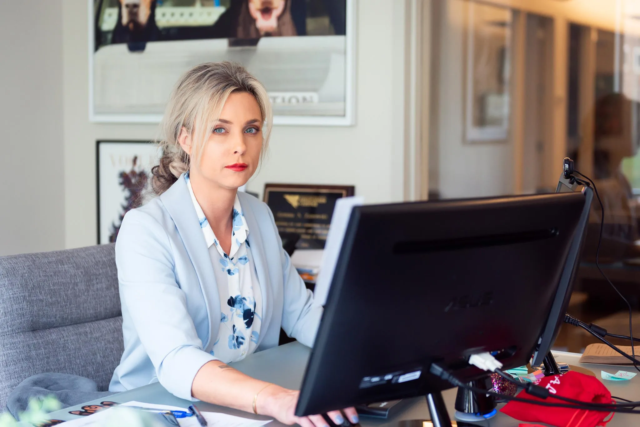 A blonde woman in a light blue blazer sits at her office desk and looks at the camera.