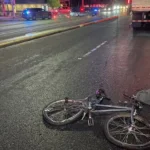 "Damaged bicycle on road at night near truck with police vehicles in background"
