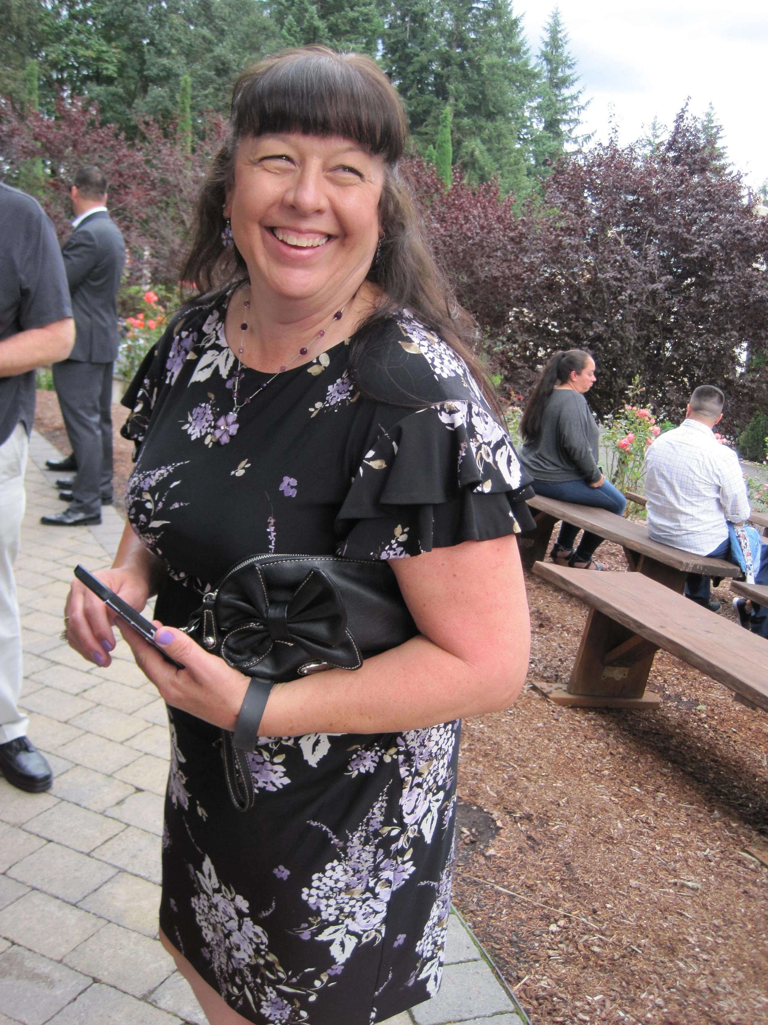 "Smiling woman in a floral dress holding a clutch outdoors at a gathering"