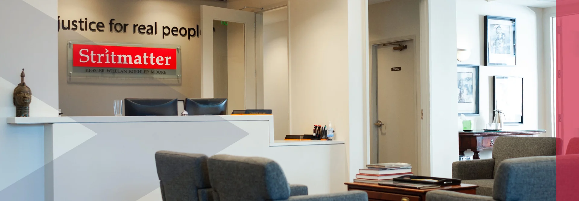 This image shows the reception area of **Stritmatter Kessler Whelan Koehler Moore**, a law firm. The office space includes a front desk with computer monitors, comfortable gray seating in the waiting area, and framed artwork on the walls. The firm’s motto, *“justice for real people”*, is prominently displayed on the wall above the red **Stritmatter** sign.
