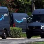 A row of dark blue Amazon Prime delivery vans, including several Mercedes-Benz Sprinter models, parked on a street lined with greenery.