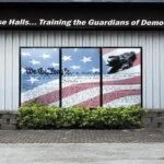 Building exterior with a patriotic mural on the windows, featuring the U.S. Constitution, American flag, and a statue of a soldier, with the caption above reading, "In These Halls... Training the Guardians of Democracy."