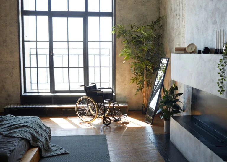 A wheelchair sits in a sunlit, modern room in front of a large window and a mirror.
