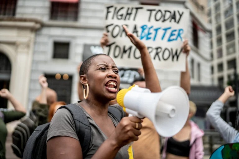 A woman yells into a megaphone at a protest, with a sign in the background.