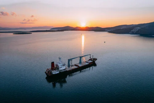 A cargo ship floating in calm waters during a sunset, with the sun setting behind distant hills and its reflection shimmering on the water.