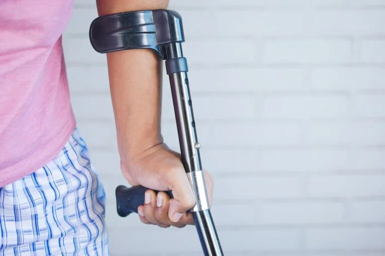 A close-up of a person's hand holding a crutch, with a white brick wall in the background.