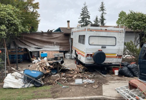 Cluttered yard with an RV, scattered debris, wood pieces, and tools; wheelbarrow in foreground and trees surrounding the area.