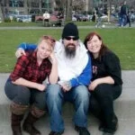 A man with a long beard sits on a ledge between two smiling women in a park.