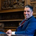 A smiling man in a blue suit sits at a table in an ornate, wood-paneled room.