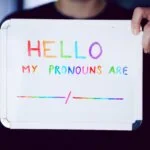 A person holds a whiteboard that reads, "Hello, my pronouns are" with a blank line.