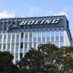 Exterior view of the Boeing corporate building with the Boeing logo prominently displayed on the upper part of the structure, framed by trees in the foreground against a clear blue sky.