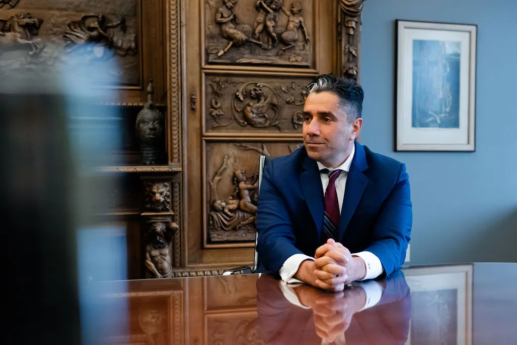 This image shows a man in a navy blue suit, white shirt, and maroon striped tie sitting at a polished wooden table. He is leaning slightly forward with his hands clasped, appearing thoughtful and engaged. The background features an ornate carved wooden panel with artistic details, and a framed picture hangs on the wall to the right. The setting looks professional, possibly an office or a conference room, conveying a formal and serious atmosphere.