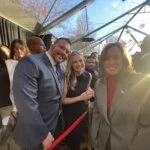 This photo shows a group of people at what appears to be a formal or celebratory event under a clear tent with sunlight streaming in. A man in a blue suit and a woman in a black dress are smiling for the camera, standing next to another woman in a gray suit with a red top. The atmosphere looks festive and warm, with many people gathered around, some holding drinks and others taking photos. The overall mood is cheerful and celebratory.