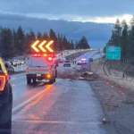 This image shows a traffic incident scene on a highway. Police and incident response vehicles with flashing lights are present, directing traffic around the obstruction. A large debris field, including broken wood and other materials, is scattered across the road. Traffic is backed up in the opposite lanes, and a green road sign indicates directions toward the Spokane Airport and Sunset Hwy exit.