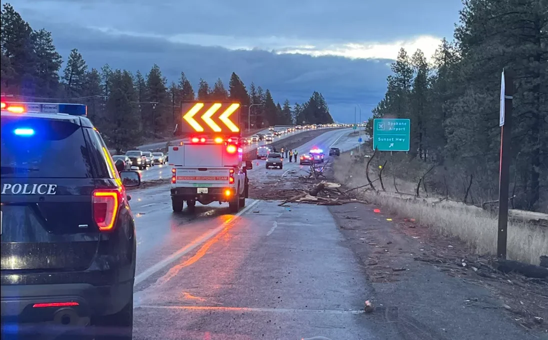 This image shows a traffic incident scene on a highway. Police and incident response vehicles with flashing lights are present, directing traffic around the obstruction. A large debris field, including broken wood and other materials, is scattered across the road. Traffic is backed up in the opposite lanes, and a green road sign indicates directions toward the Spokane Airport and Sunset Hwy exit.