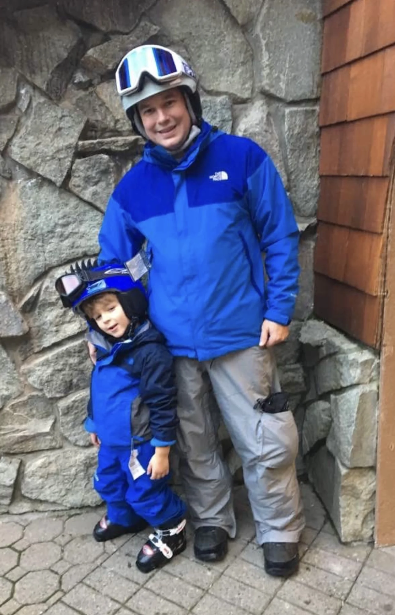 "Man and child in blue ski gear posing together with helmets and goggles."