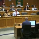 "Two people testify before a government panel in a formal hearing room."