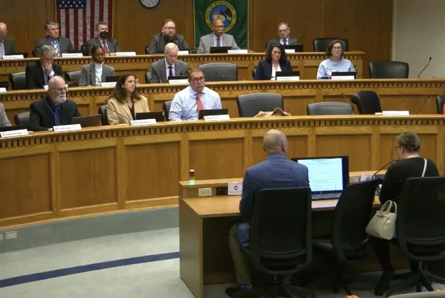 "Two people testify before a government panel in a formal hearing room."