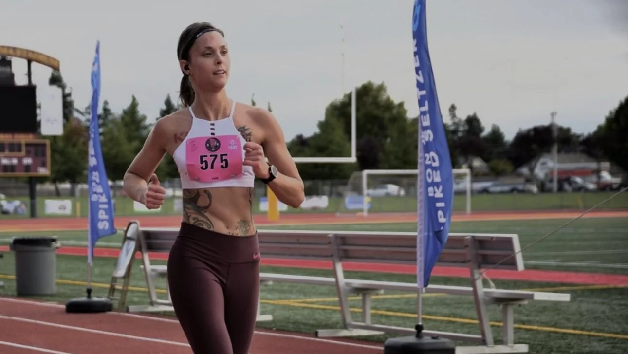 Woman running on a track during a race, wearing bib number 575, maroon leggings, and a white sports bra, with sponsor flags and stadium in the background