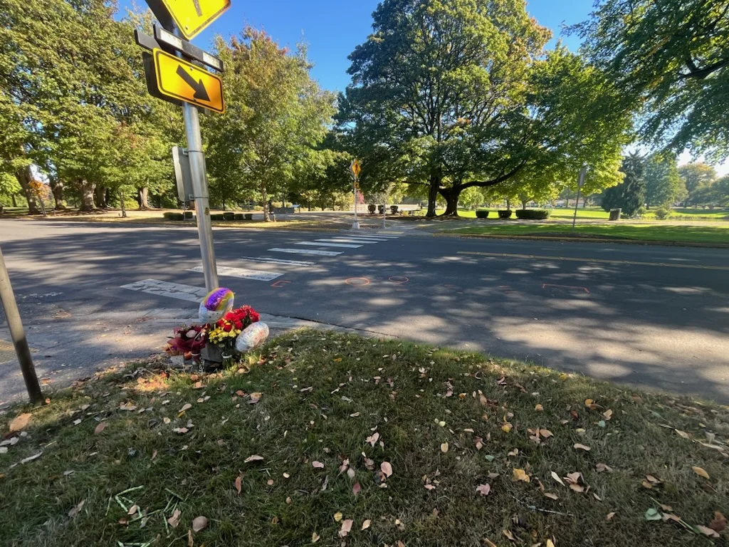 "Roadside memorial with flowers and balloons near a crosswalk under trees."