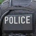 Close-up of the back of a police officer wearing a tactical vest with a large "POLICE" label and a coiled radio wire.