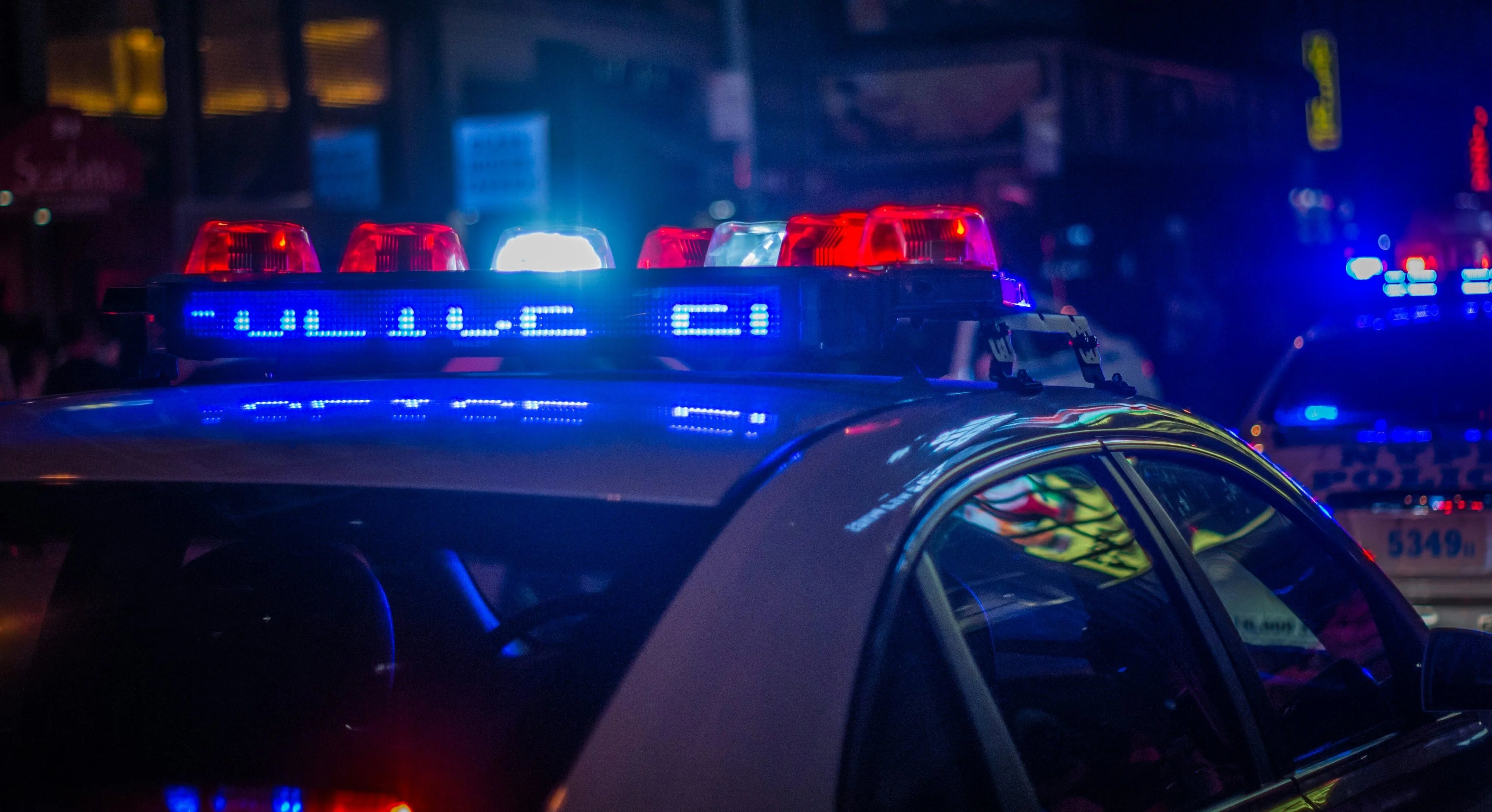 Close-up of a police car with flashing red and blue lights at night in an urban setting, reflecting city lights and surrounded by other emergency vehicles.