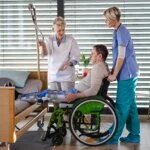 A male patient with leg braces sits in a green wheelchair next to a hospital bed, assisted by two female healthcare professionals.