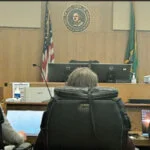 Three attorneys seated at the counsel table in a courtroom, facing the judge’s bench with the Washington State seal, American flag, and Washington State flag visible in the background.