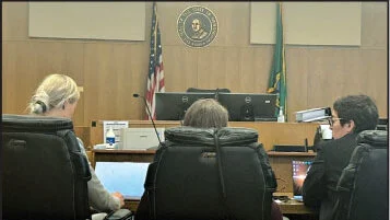 Three attorneys seated at the counsel table in a courtroom, facing the judge’s bench with the Washington State seal, American flag, and Washington State flag visible in the background.