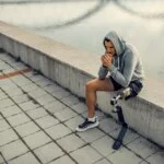 Man with a prosthetic leg sitting on a waterfront ledge, looking thoughtful.