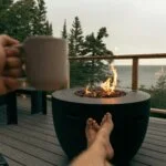 Person relaxing with a drink near a modern outdoor fire pit on a rainy deck overlooking the water.
