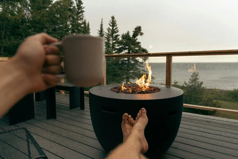 Person relaxing with a drink near a modern outdoor fire pit on a rainy deck overlooking the water.