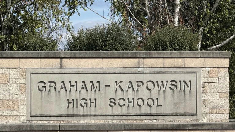 Stone sign reading 'GRAHAM-KAPOWSIN HIGH SCHOOL' surrounded by trees and bushes.