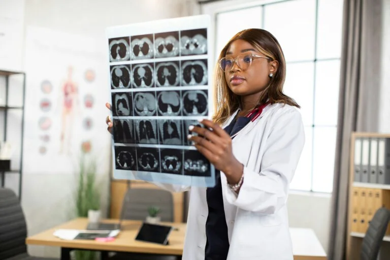 A medical professional wearing a white lab coat holds up a large sheet of brain scan images in a bright office setting.